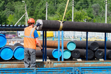 Slinger in hard hat and high-visibility vest unloads metal pipes for gasification on summer day in...