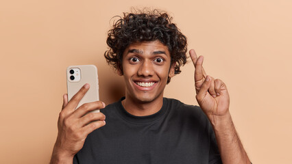 Studio shot of curly haired Hindu man smiles toothily holds cellphone and keeps fingers crossed believes in good luck hopes for good fortune wears black t shirt isoalted over brown background