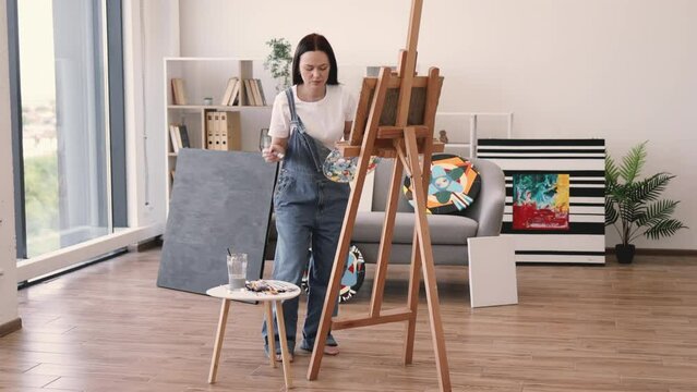 Full length view of barefoot woman in denim overalls working on easel in spacious art studio on sunny day. Confident adult focusing on paint application using wide brush in comfortable interior.