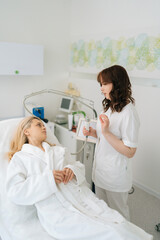 Vertical shot of female cosmetologist consulting woman client in white bathrobe lying on medical couch before face rf-lifting procedure. Concept of non-surgery cosmetology, professional skincare