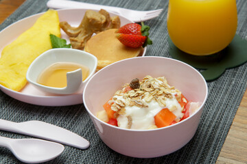 Kids menu - omlet, pancakes with maple syrup, muesli with yoghurt and glass of fresh squuzed orange juice served on the restaurant table with plastic kids friendly eating utensil.