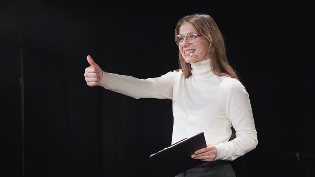 Female conference presenter in headset doing happy thumbs up gesture during motivational speech on stage. Charming caucasian woman holding clipboard and sincerely smiling.
