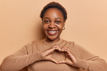 Pretty young African woman exudes pure happiness as she smiles and forms heart gesture with both hands smiles broadly dressed in casual jumper isolated over brown background. Body language concept