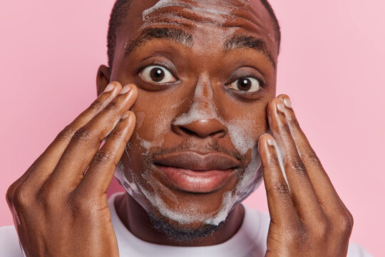 Close up shot of dark skinned surprised young man keeping palms on cheeks undergoes hygiene procedures has bugged eyes applies foaming bubble isolated over pink background. Cleanliness concept