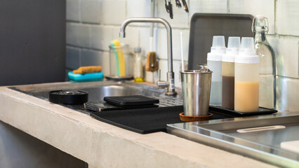 A stainless steel cup placed on kitchen sink with set of coffee making ingredient such as condensed milk and sweet syrup bottles. 