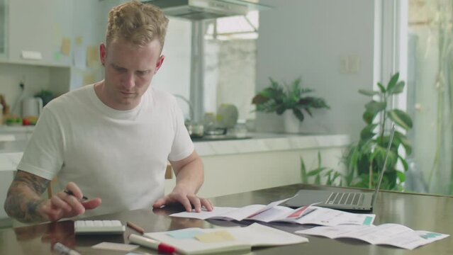 Medium shot of young man sitting at home, checking printed bills and using calculator while planning his budget