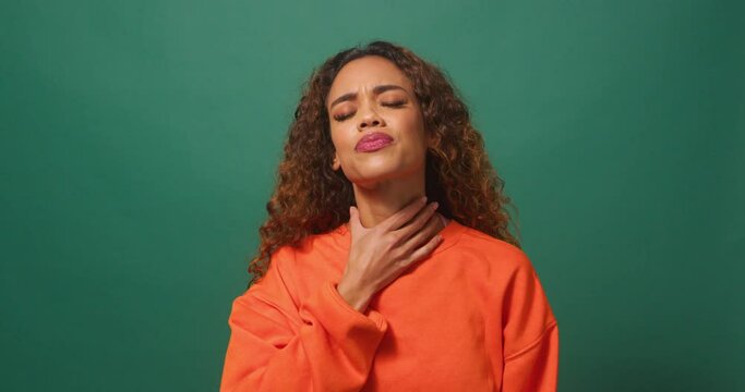 Young multi-ethnic woman rubs sore throat in pain, green background studio
