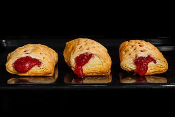 Three baked puff pastry buns with red strawberry jam on tray in electric oven, black background - front view. Homemade bakery, food, cooking and pastry concept