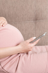 pregnant woman using her smartphone touching her belly sitting on couch. woman texting message on her phone
