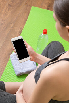 Pregnant female holding smartphone while sitting on exercise yoga mat at home at coronavirus time