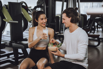 Young beautiful woman and handsome boy sitting drinking milk, whey protein and healthy, clean food to maintain health against backdrop of exercise equipment in gym