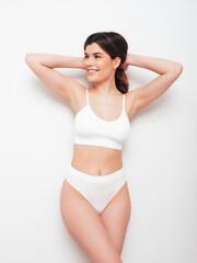 Obraz premium Portrait of young beautiful sexy woman in white lingerie. Smiling carefree model wearing pure underwear. Hot brunette posing near wall in studio interior. Perfect body. Slim and fit