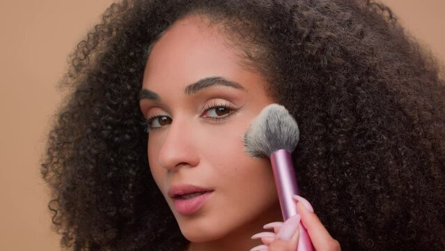 Close up portrait at beige studio background beautiful African American woman attractive ethnic girl looking at camera apply powder for face make-up preparing cosmetics beauty blush bronzer contouring