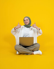 Showing thumbs up, full body young muslim woman wear hijab showing thumbs up. Pretty woman in head scarf sitting ground notebook on her lap. Using computer, office worker, business woman. Lifestyle.