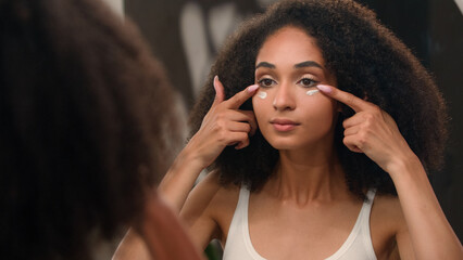 Ethnic girl beauty routine in morning young African American woman applying face cream under eyes anti-aging skin care dark eye circles moisturizing organic natural cosmetic facial mask looking mirror