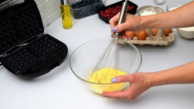 Woman chef beating eggs into bowl,mixing with whisk,cooking dough for baking waffles,pancakes.Morning breakfast food preparation,recipe step instruction on kitchen table.healthy eating,bakery
