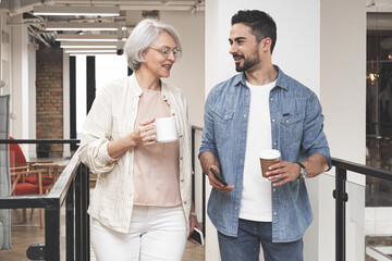 Smiling adult woman and young man walking in the office while talking, holding coffee. Startup, coworking, management concept