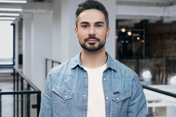 Naklejka premium Portrait of young bearded man standing in the office while posing at camera. Modern coworking on the background. Startup, coworking, management concept