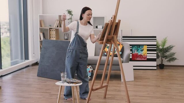 Young beautiful female smiling while making selfie on personal gadget in stylish apartment with large panoramic windows. Creative artist posing near big wooden easel and small table with paints.