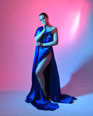 Full length portrait of beautiful female model wearing elegant flowing toga gown of blue silk drapery, isolated on studio background with cinematic colourful lighting.