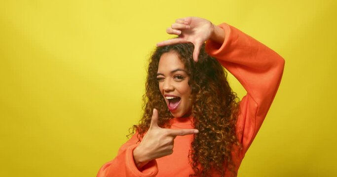 Young multiracial woman frames face with rectangle fingers, yellow studio