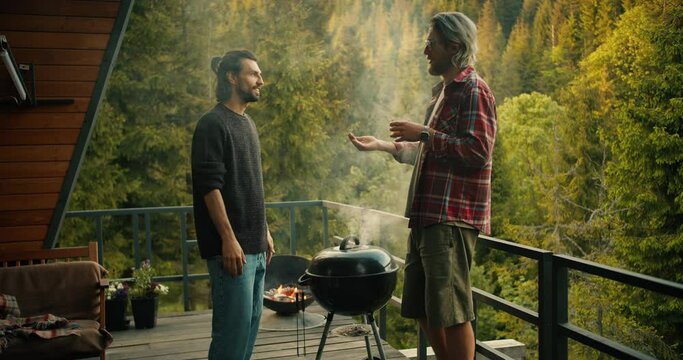 A couple of friends chat while cooking on the grill. A brunette guy in a green sweater lifts the lid of the grill and thick white smoke comes out. Picnic with a view of the mountains and coniferous