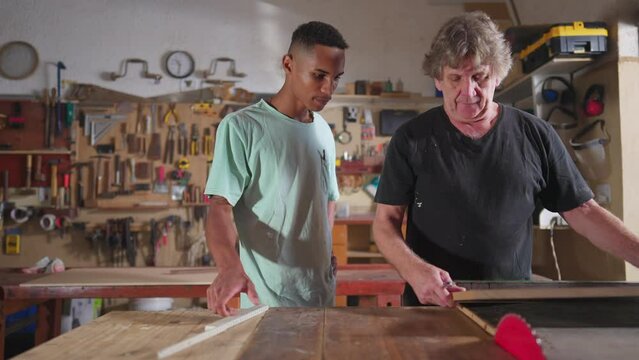 Carpenter and apprentice working at workshop, teacher orienting young man with saw machine at carpentry. Wood sawing scene of mentor teaching student