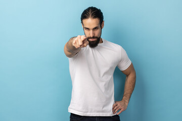 Portrait of serious man with beard wearing white T-shirt looking at camera and blaming warning you with raised finger, showing admonishing gesture. Indoor studio shot isolated on blue background.