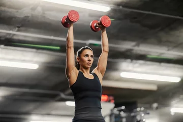 Fotobehang Sportschool A muscular sportswoman in shape is lifting dumbbells over her head in a gym.  © Dusan Petkovic