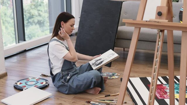 Side view of focused female painter wearing denim jumpsuit sitting on wooden floor and working on new drawing at own art studio. Young lady spending free time with canvas and paintbrush in hands.