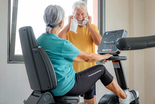 Athletic senior couple stands together in gym, woman doing exercise bike while talks with elderly man