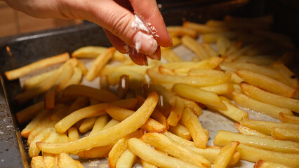 Closeup of female hand pouring salt on frying french fries potatoes on baking pan. Fast food, healthy nutrition, cooking in oven.