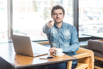 Fototapeta na wymiar Portrait of strict bossy young bearded handsome man freelancer in blue jeans shirt working on laptop, pointing at camera, selecting you. Indoor shot near big window, cafe background.