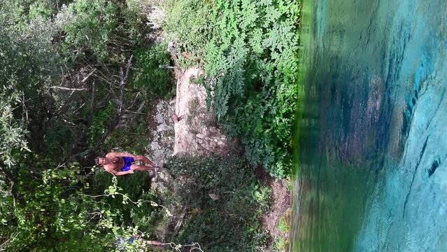 Tourist man jump into unique karst source of water gushing from the ground called the Blue Eye or Syri i Kalter. Muzine, Albania. River framed by green trees. Summer landscape.