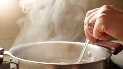 Female hand stirring boiling soup in metal pan with spoon. Vapour slowly rising from hot water.