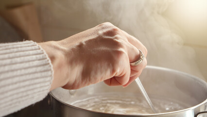 Closeup of woman stirring ingredients in pot while cooking soup. Healthy nutrition, cooking at home, hot steam.