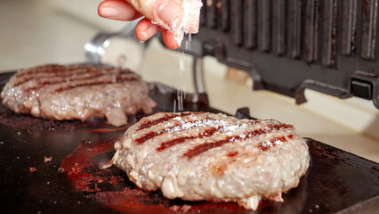 Closeup of hand salting beef burgers cooking on electric grill at home. Cooking at home, kitchen appliance, healthy nutrition, hamburger ingredients.