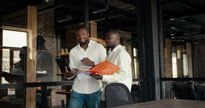 Two Black-skinned businessmen in white shirts are walking around the loft-style office and discussing new projects using a laptop and a folder with documents