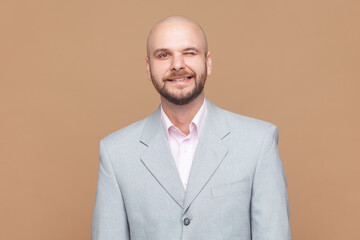 Portrait of playful positive optimistic bald bearded man has fun and winks eye, smiles with teeth makes funny grimace, wearing gray jacket. Indoor studio shot isolated on brown background.