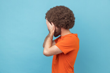 Side view of man with Afro hairstyle in orange T-shirt closing eyes with hand, dont want to see that ignoring problems, hiding from stressful situations. Indoor studio shot isolated on blue background - obrazy, fototapety, plakaty