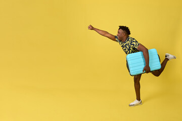 Young African man in a superman pose holds a blue travel suitcase on a yellow background. Happy man...