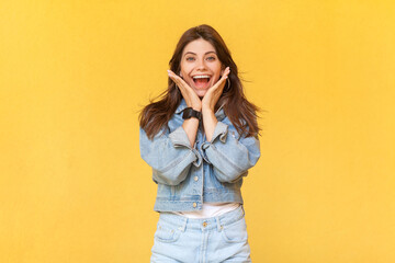 Portrait of excited extremely happy woman in denim jacket standing with palms near face, screaming happily, rejoicing her victory, screaming hurray. Indoor studio shot isolated on yellow background.