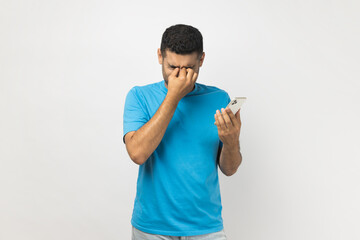 Portrait of tired sad unshaven man wearing blue T- shirt standing holding smart phone in hands,...