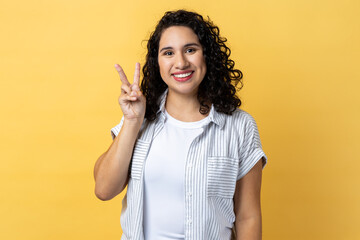 Portrait of happy optimistic woman with dark wavy hair doing victory gesture and looking smiling to...
