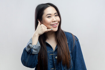 Fototapeta na wymiar Give me tell telephone number. Portrait of brunette woman in blue denim jacket standing makes call me back gesture, has optimistic face expression. Indoor studio shot isolated on gray background.