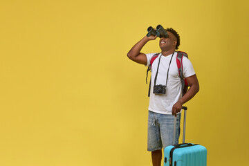 Positive black tourist with a suitcase and backpack stopped looking at the image through binoculars. A traveler with luggage in fashionable summer clothes went on vacation with binoculars