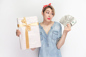 Portrait of confused puzzled uncertain blonde woman wearing blue denim shirt and red headband standing holding present box and dollar banknotes. Indoor studio shot isolated on gray background.