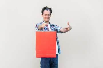 Man holding and pointing at bag with purchases, being in high spirits, spending free time in shopping mall, wearing blue checkered shirt and headband. Indoor studio shot isolated on gray background.