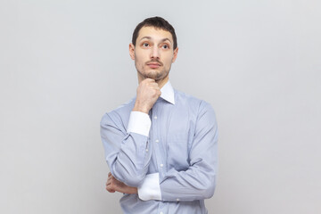 Portrait of pensive puzzled young adult man holding chin, thinking about his work, looking away with thoughtful expression, wearing light blue shirt. Indoor studio shot isolated on gray background.