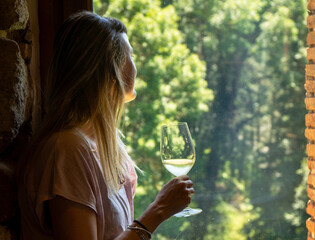 Young, pretty woman with long blonde hair looking through a window holding a glass of white wine in...